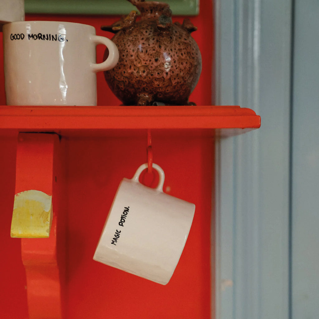 White mug with 'Magic Potion' text on a red shelf with a decorative vase and another white mug.
Magic Potion Mug - Jo And Co Magic Potion Mug - Anna + Nina